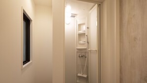 Un couloir étroit mène à un espace douche, avec une fenêtre sur la gauche et une porte en bois sur la droite.