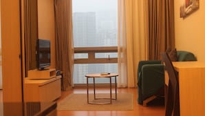 Ett hotellrum med en tv på ett träställ, ett litet runt bord, en grön stol och ett stort fönster med gardiner.