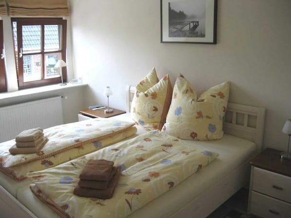 Una cama bien hecha con ropa de cama floral y varias almohadas, una mesita de noche con lámpara y un cuadro en la pared.
