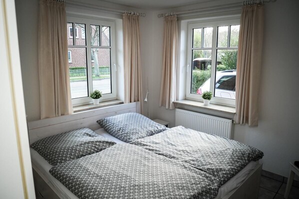 Une chambre avec deux lits, des rideaux de couleur beige, une fenêtre donnant sur la rue et une petite plante posée sur le rebord de la fenêtre.