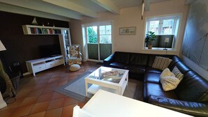 A living room with a black leather sofa, a white coffee table, a floral armchair, a bookshelf, and a television. - Boowen Taarep 19, Germany (Utersum)