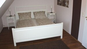 A bedroom with a white bed, two bedside tables, a nightstand, a wall-mounted lamp, and a framed picture. - Boowen Taarep 19, Germany (Utersum)