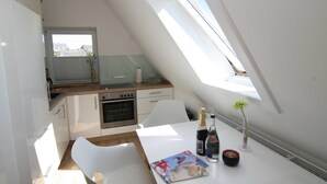 A modern kitchen with white cabinetry, a wooden countertop, and a built-in oven. There is a dining table with a magazine, a bottle of champagne, and a vase with flowers. A window provides natural light and a view of buildings outside. - Am Wasser OG, Germany (Hörnum (Sylt))