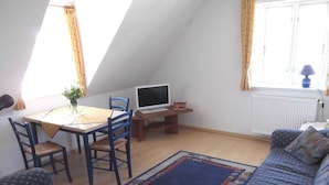 A cozy attic room with a dining table, chairs, a TV, a sofa, and a patterned rug.