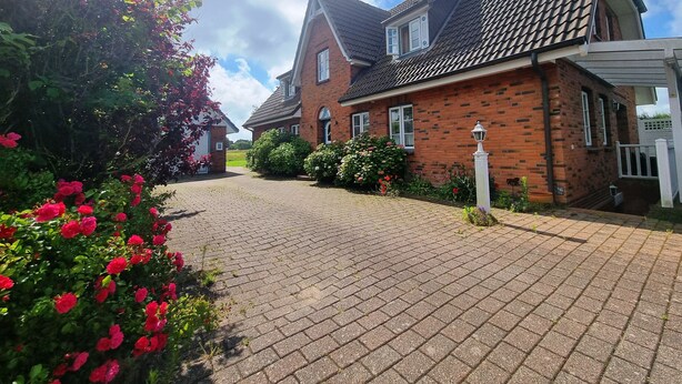 A brick house with a tiled roof, a paved driveway, and a garden with red flowers.
