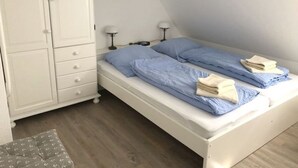 A bedroom with a single bed, a white wardrobe, two bedside lamps, and a grey patterned ottoman. - Neiwoi 2c, OG, Germany (Borgsum)