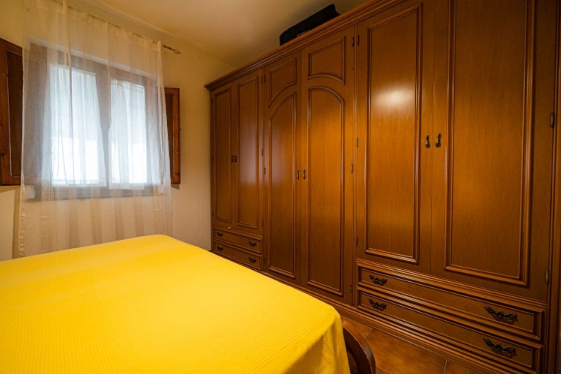 A room with a bed, wooden wardrobe, and a window with sheer curtains.