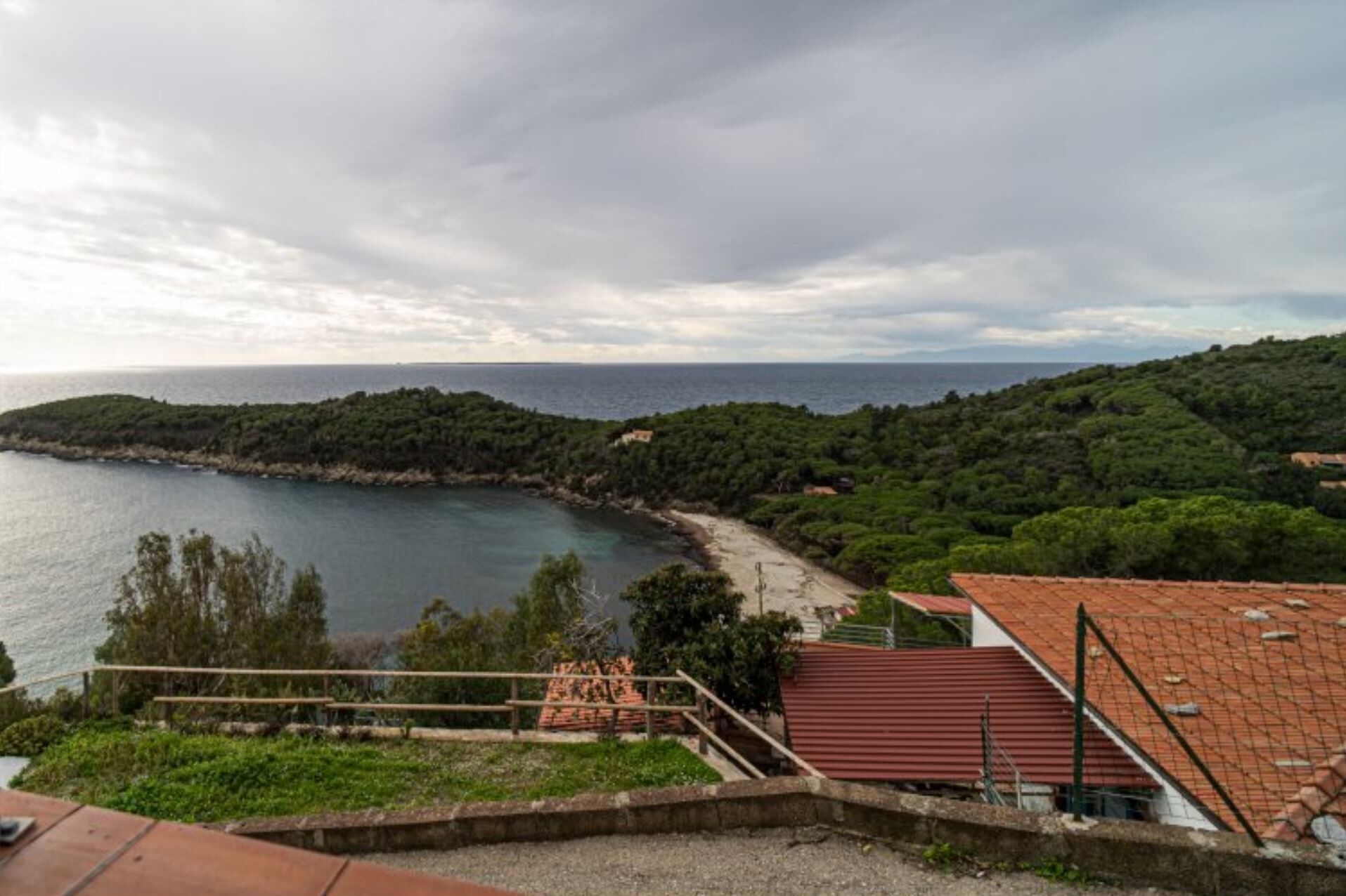 En kustvy med en strand, en trappa som leder upp till en byggnad med ett rött tak och en skogbeklädd sluttning.