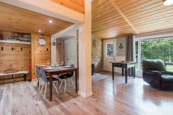 A spacious room with a wooden ceiling, a dining area with a table and chairs, a foosball table, and a view of the outdoors.