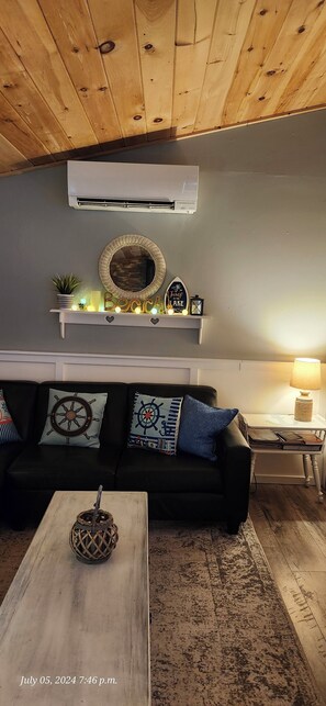 Een woonkamer met een houten plafond, een zwarte bank met marinekussens, een witte plank met decoratieve objecten en een houten salontafel.