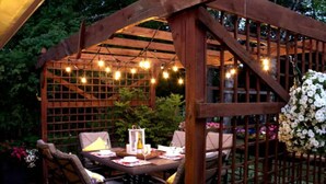 En pergola av trä med ljusslingor, ett bord med stolar och en blomsterdekoration.
