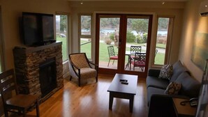 Una sala de estar con chimenea de piedra, un televisor montado encima, un sofá, una mesa de centro y vistas a un jardín a través de puertas de vidrio.