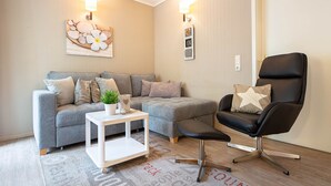 Un salón con un sofá gris, una silla negra, una mesa de centro blanca y una alfombra estampada.