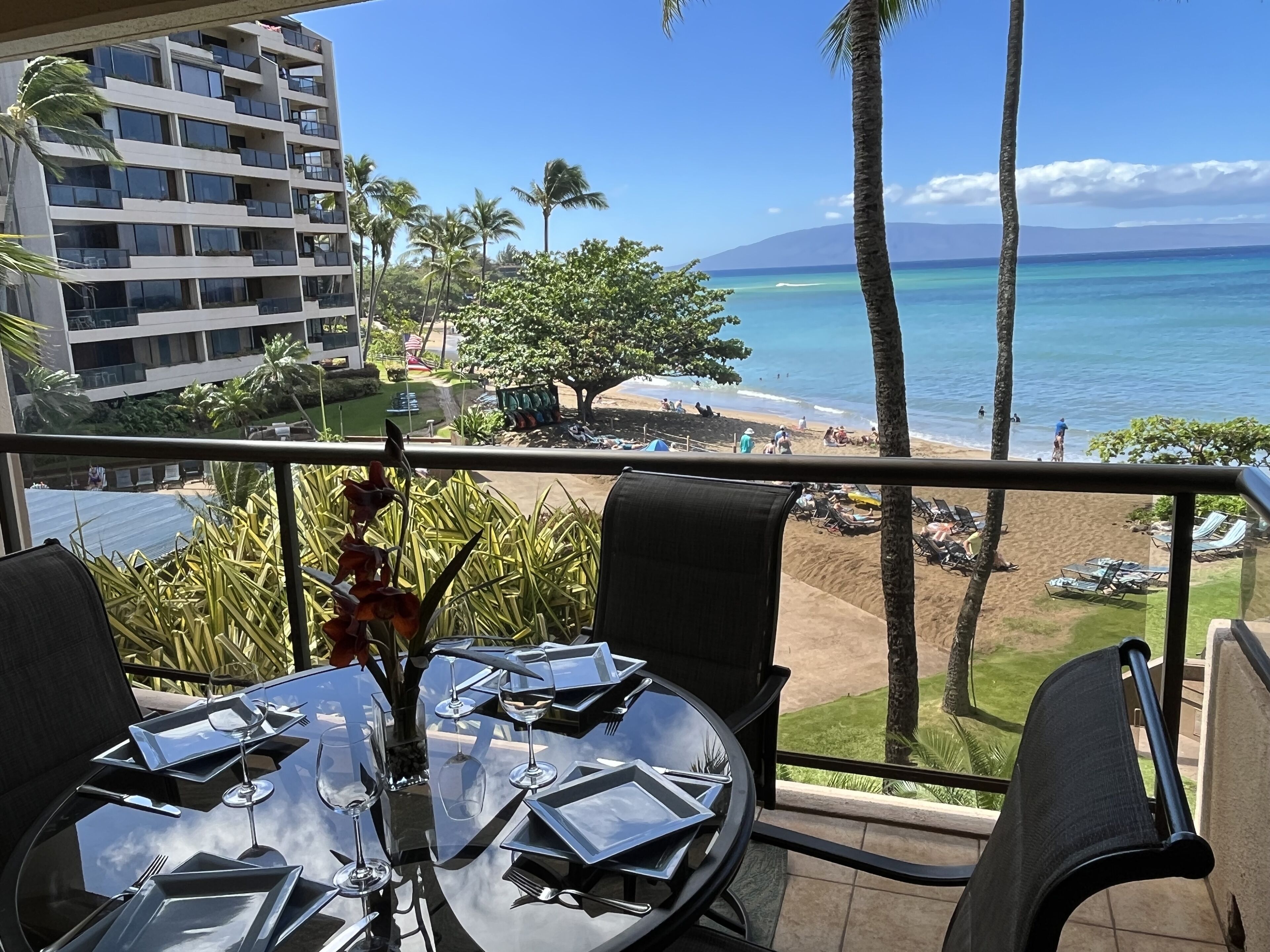 A balcony with a table set for dining, overlooking a beach with palm trees and a view of the ocean.
