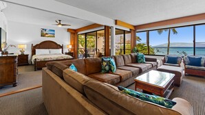 Un salón amplio con un sofá grande color café, una mesa de centro de madera y una cama con cabecera de madera. La habitación tiene ventanas amplias que ofrecen vistas al océano y palmeras.