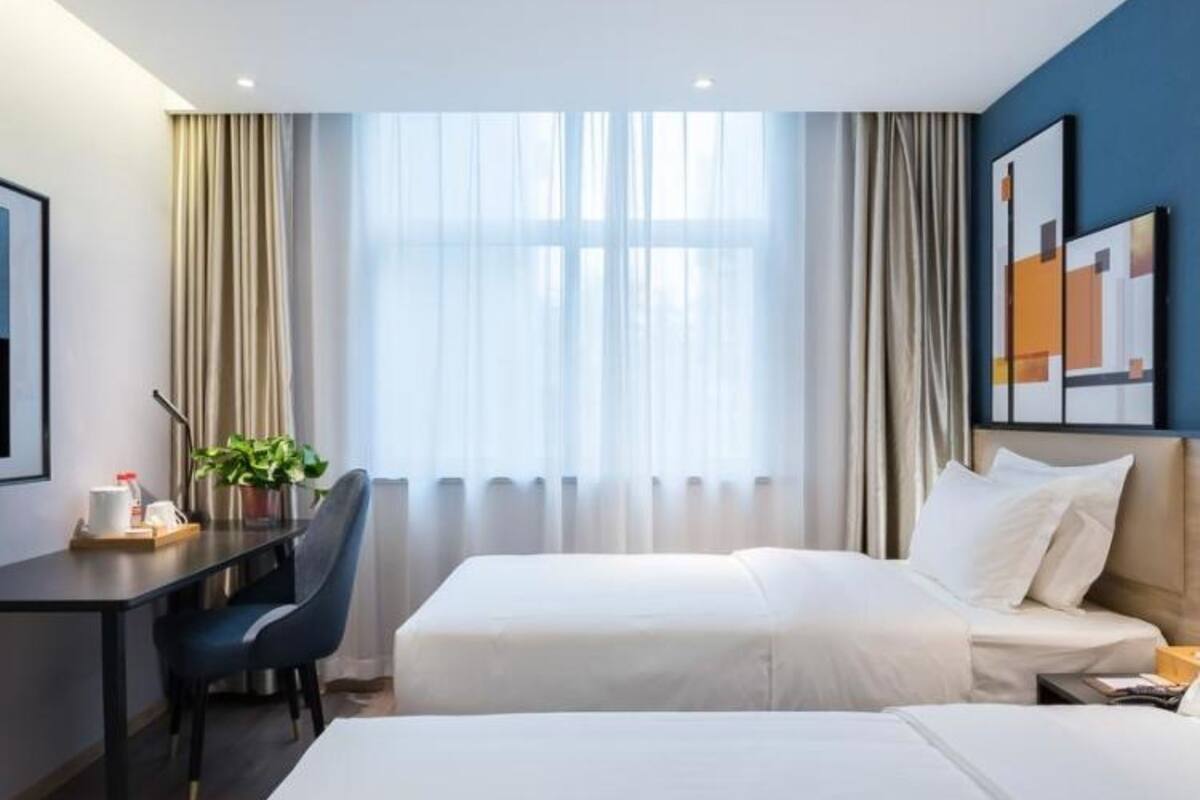 Ein Hotelzimmer mit Schreibtisch, zwei Betten, einem Sessel, einer Zimmerpflanze und einem Vorhangfenster.