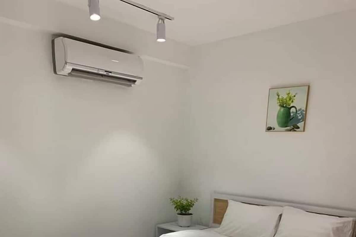 Ein ordentlich gemachtes Bett mit weißer Bettdecke in einem Zimmer mit Holzboden, Wandklimaanlage und einer kleinen Topfpflanze auf einem Nachttisch.