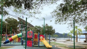 Un parque infantil con toboganes y estructuras para escalar, rodeado de árboles y un cuerpo de agua al fondo.