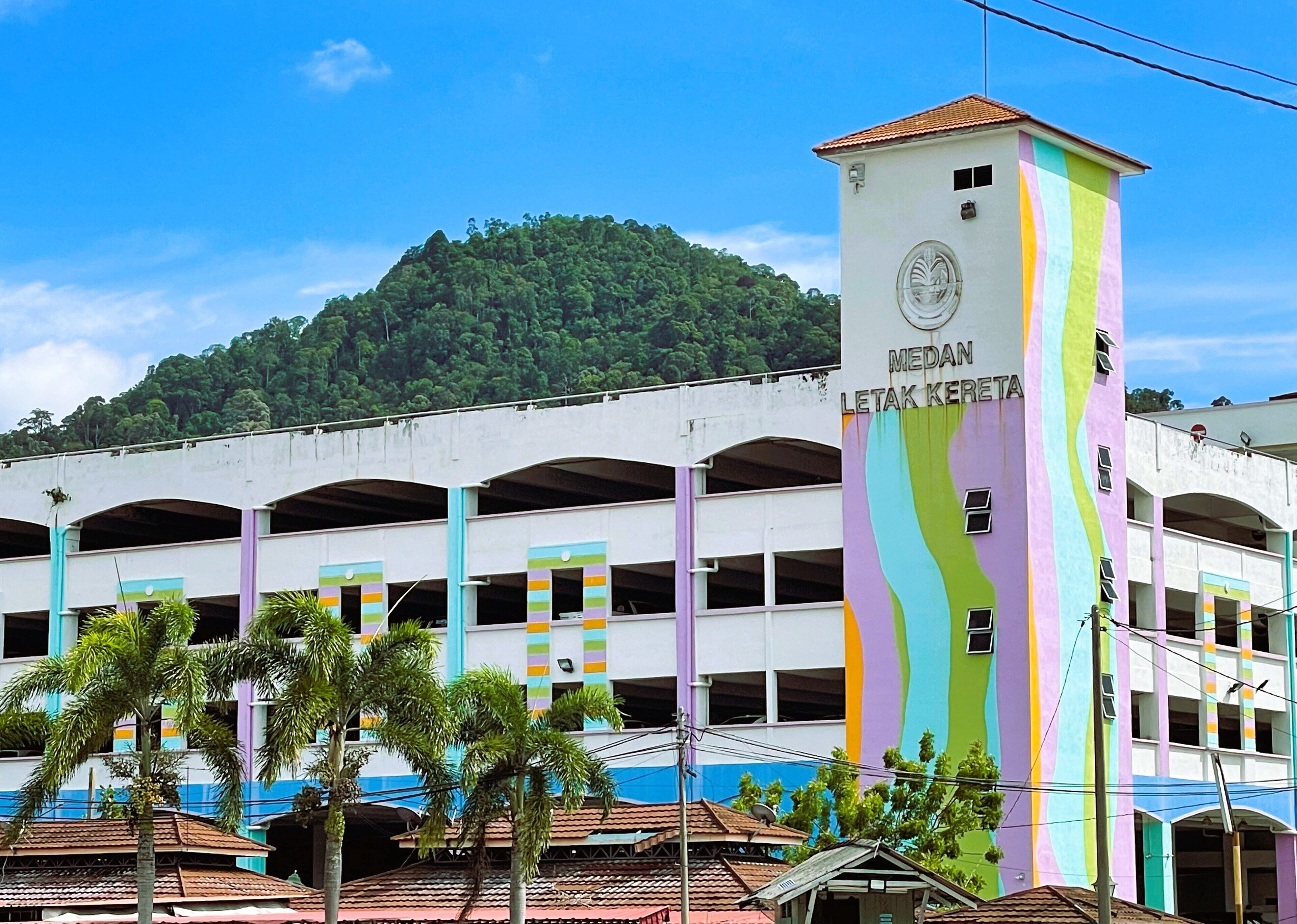 Una estructura de estacionamiento de varios pisos con rayas verticales de colores y una torre con la inscripción 'MEDAN LETAK KERETA'.
