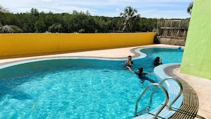 Una piscina con agua cristalina azul, rodeada por un deck con baldosas y una pared amarilla.