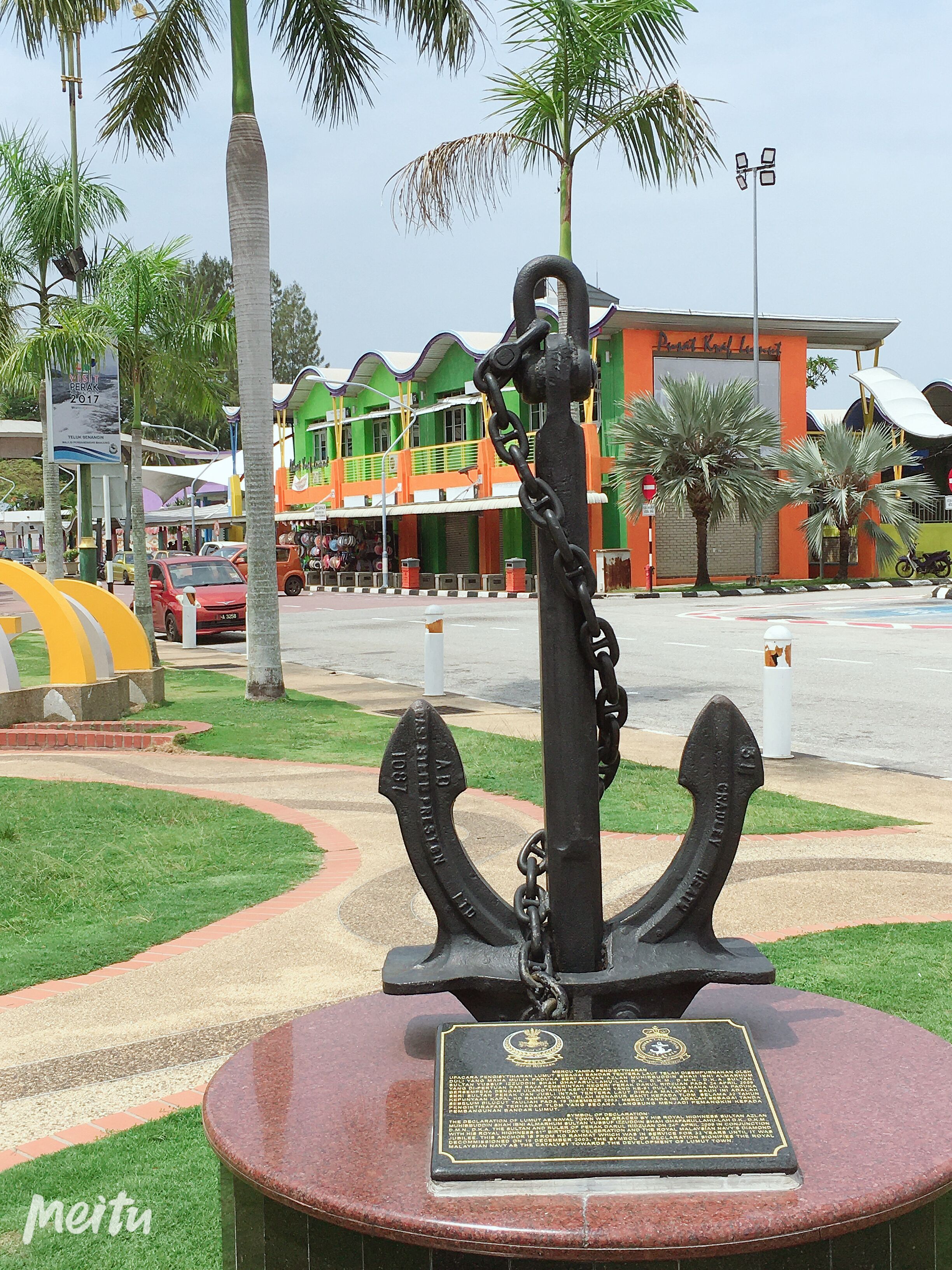 A monument with a plaque, surrounded by a circular grassy area with palm trees.
