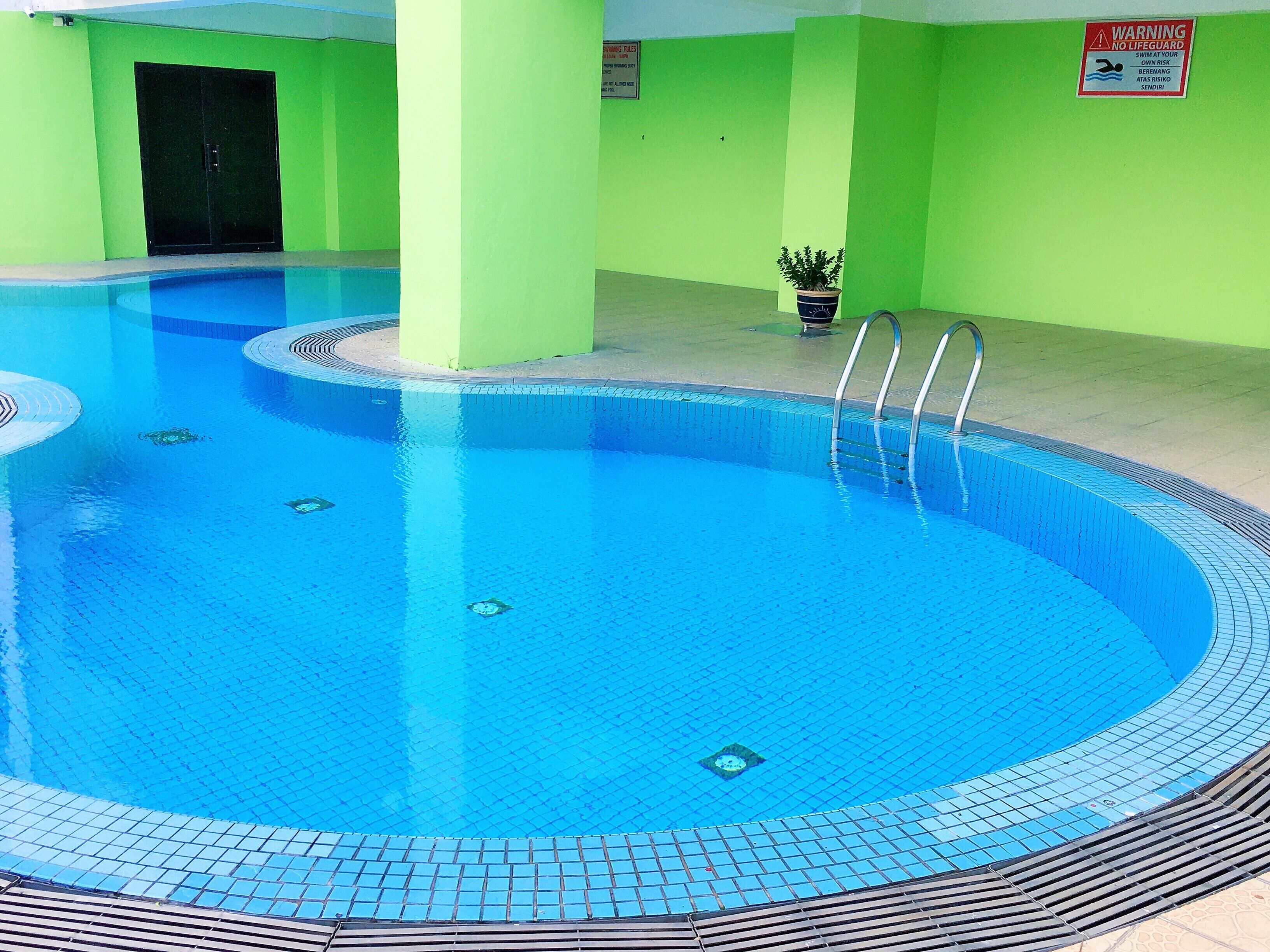 An indoor swimming pool with a tiled edge, a lifeguard warning sign, and a potted plant near the wall.