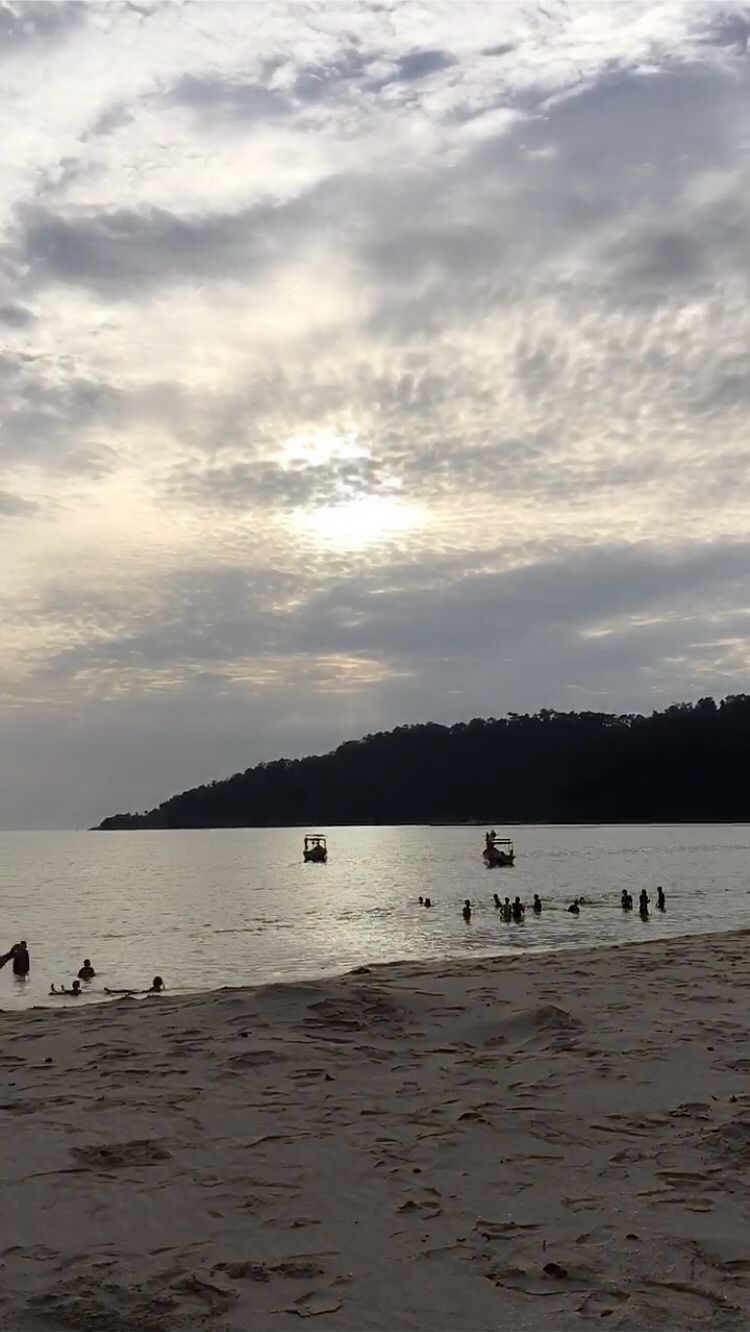Una playa con gente nadando y caminando por la orilla, barcos en el agua y un paisaje montañoso al fondo.