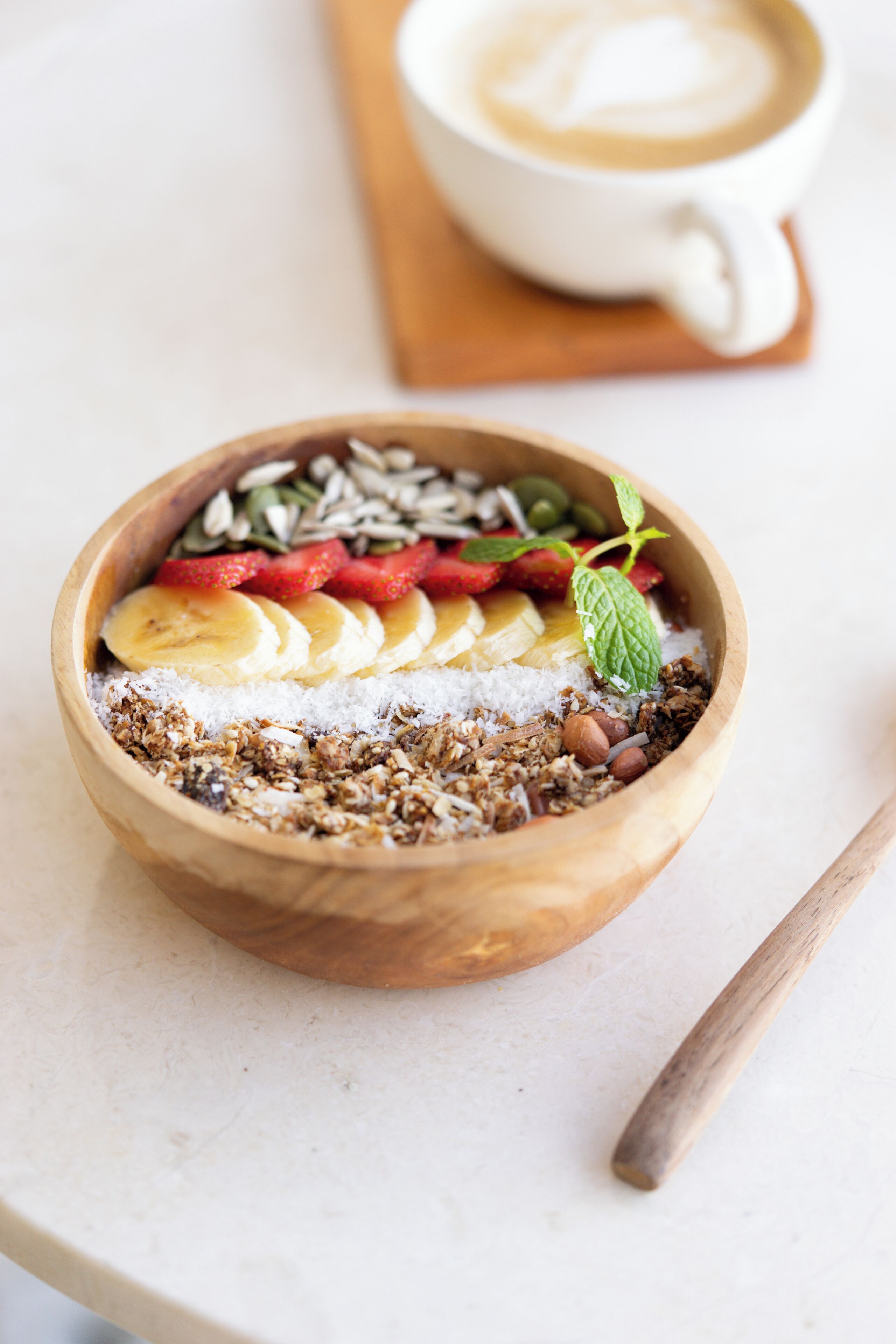 Eine Holtschale mit Granola, Bananenscheiben, Erdbeeren und Nüssen auf einer weißen Oberfläche, neben der ein Holzlöffel liegt.