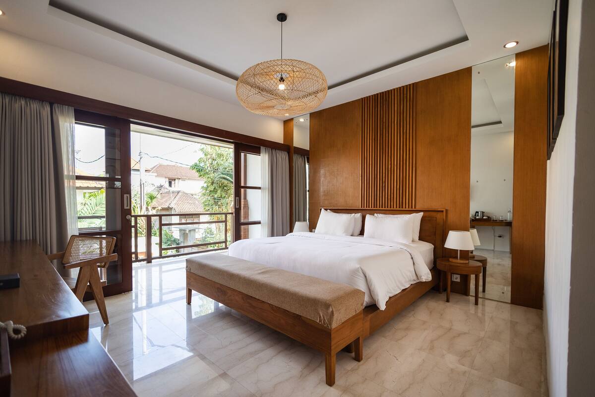 Ein modernes Hotelzimmer mit großem Bett, Holz-Kopfteil, Essbereich mit einer Bank und einem Balkon mit Blick auf Grünflächen.