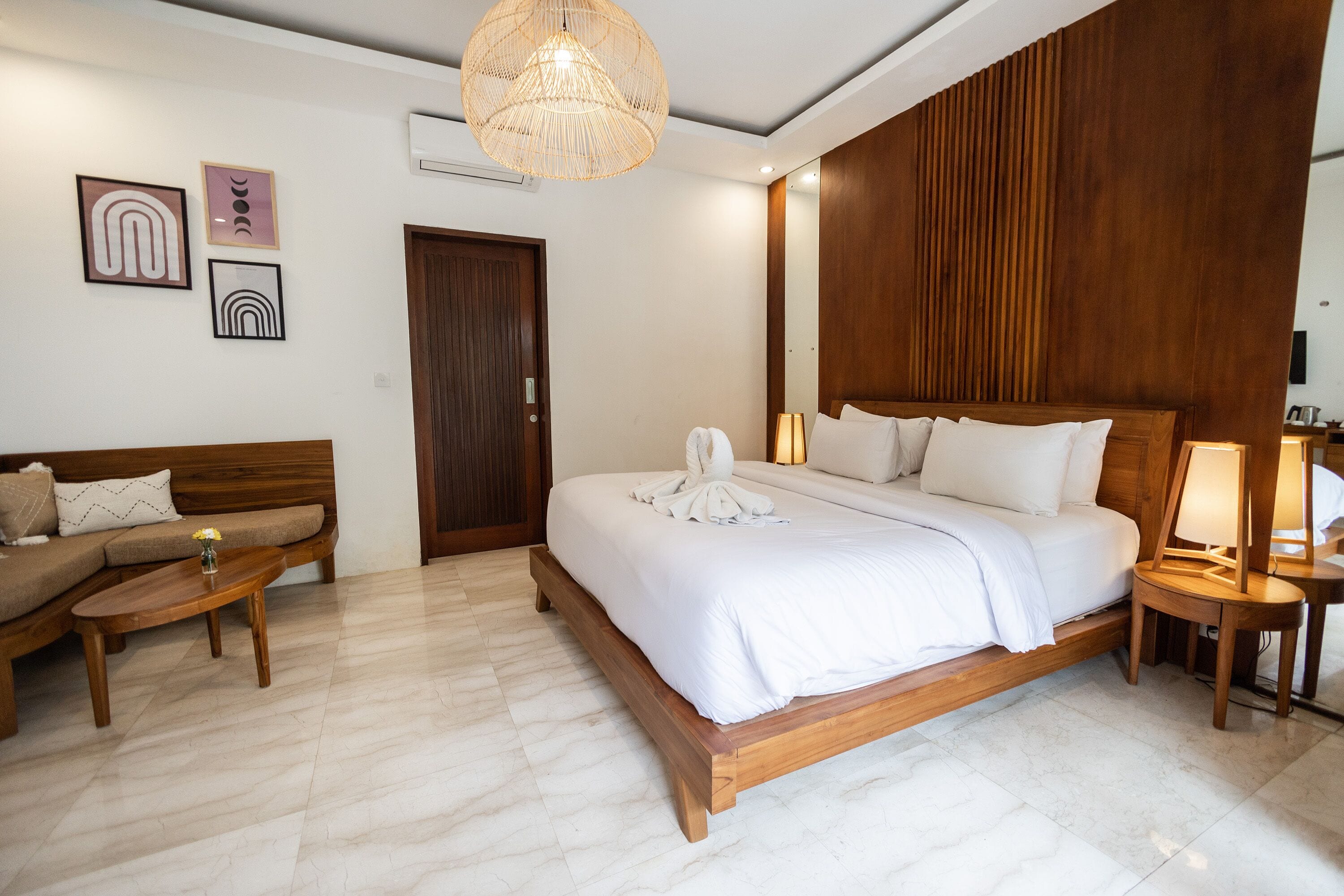 Een hotelkamer met een houten bed, een houten bankje, een tafeltje en een luchtverkoeler aan de muur.