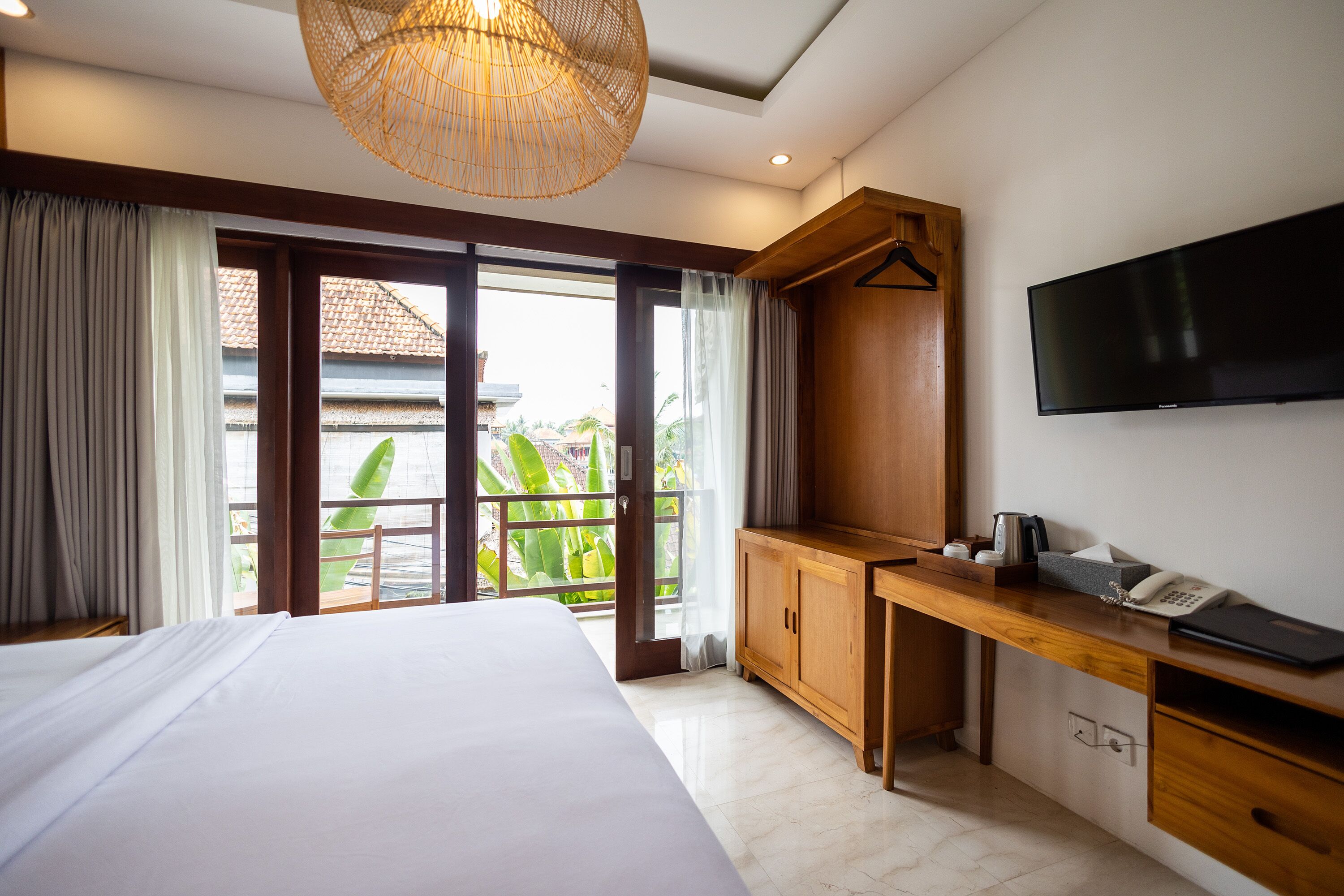 A hotel room with a large bed, a wooden desk, a television, and a balcony with a view of palm trees.