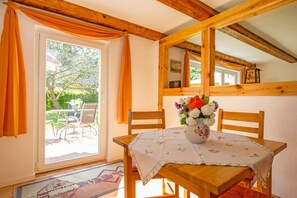 Uma mesa de jantar de madeira com toalha de mesa florida, rodeada por cadeiras de madeira, um vaso com flores e vista para um pátio ao ar livre com mesa e cadeiras.