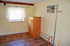 A room with wooden flooring, a window with white curtains, a wooden wardrobe, and a painting on the wall.