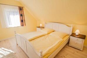 Uma cama de casal com duas mesas de cabeceira, uma janela com cortinas leves e xadrez, e piso de madeira.