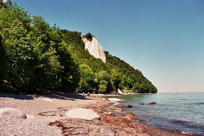 Ein Kiesstrand mit felsiger Küstenlinie, ein mit Grün bewachsener Felsen und ruhiges Wasser.