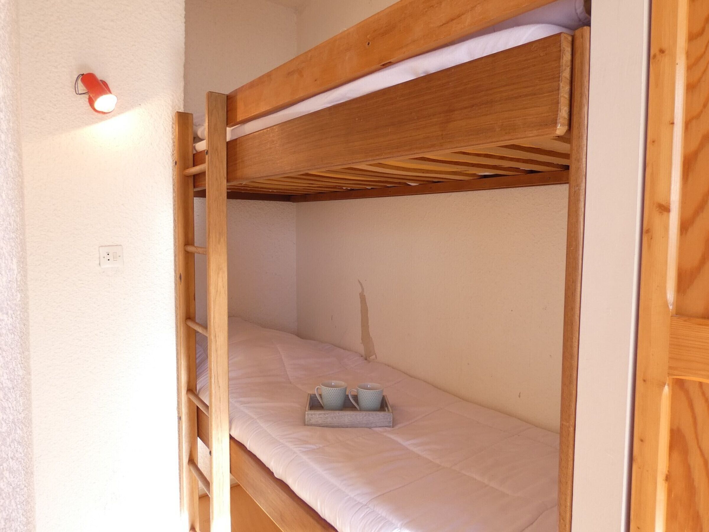 A bunk bed with a ladder, a bedside table with a cup and a bowl, and a wall-mounted light.