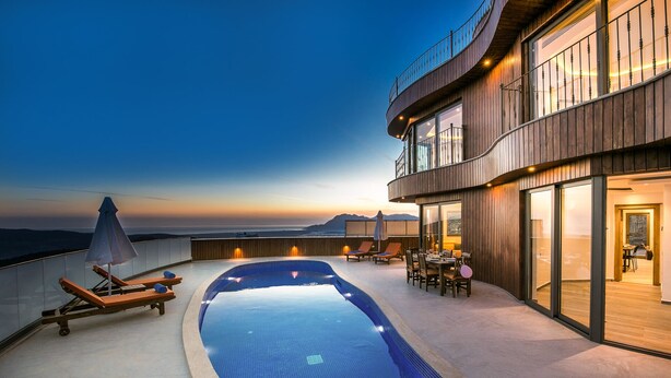 A rooftop infinity pool with lounge chairs and umbrellas, a dining area, and a view of the ocean at sunset.