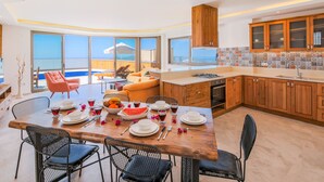 A modern kitchen with wooden cabinets, a dining area with a wooden table set for four, and a view of the ocean through large windows.