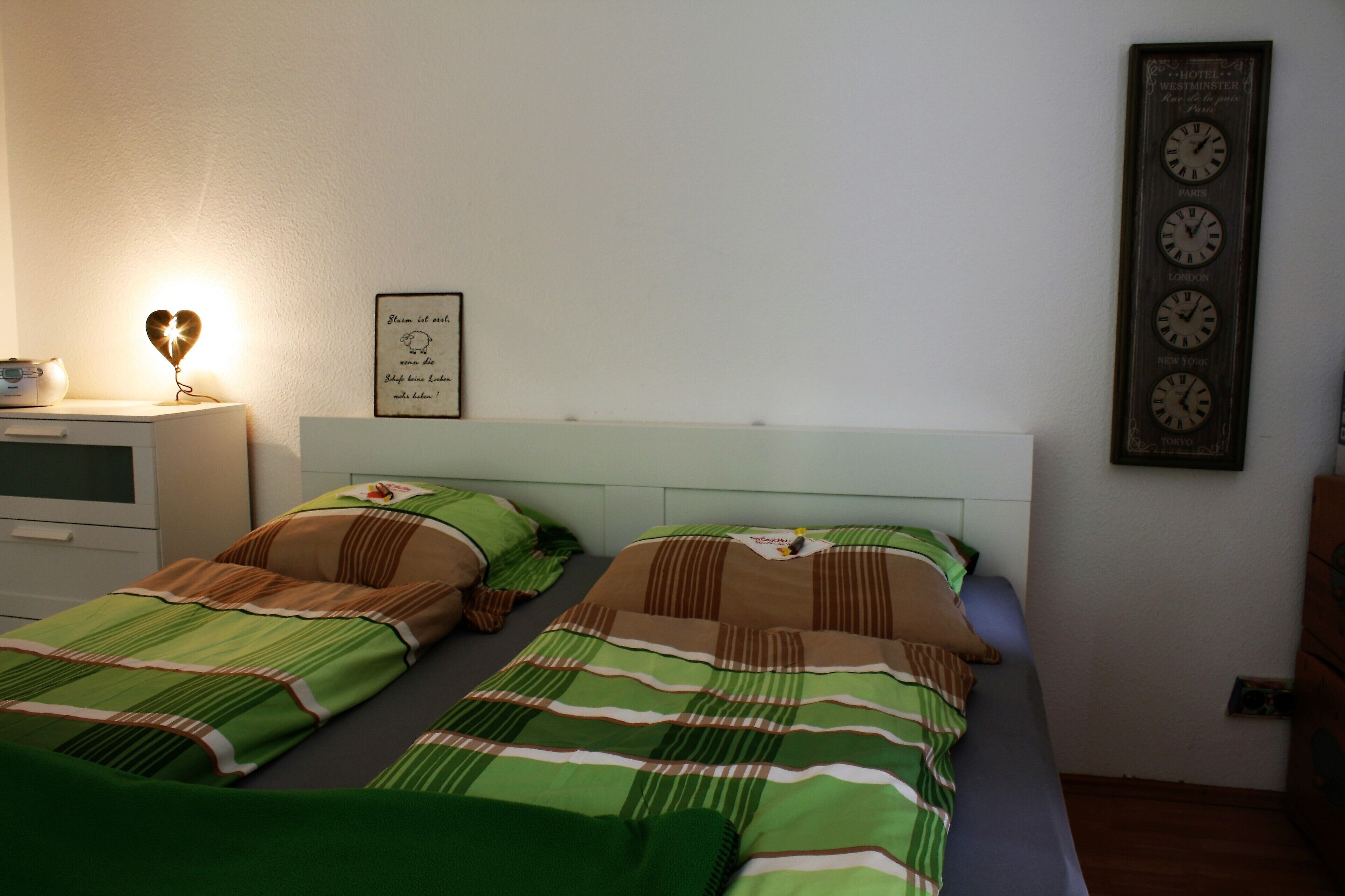 Une chambre avec deux lits simples, une table de chevet avec une lampe, une citation encadrée au mur et une horloge murale indiquant l’heure de différentes villes.