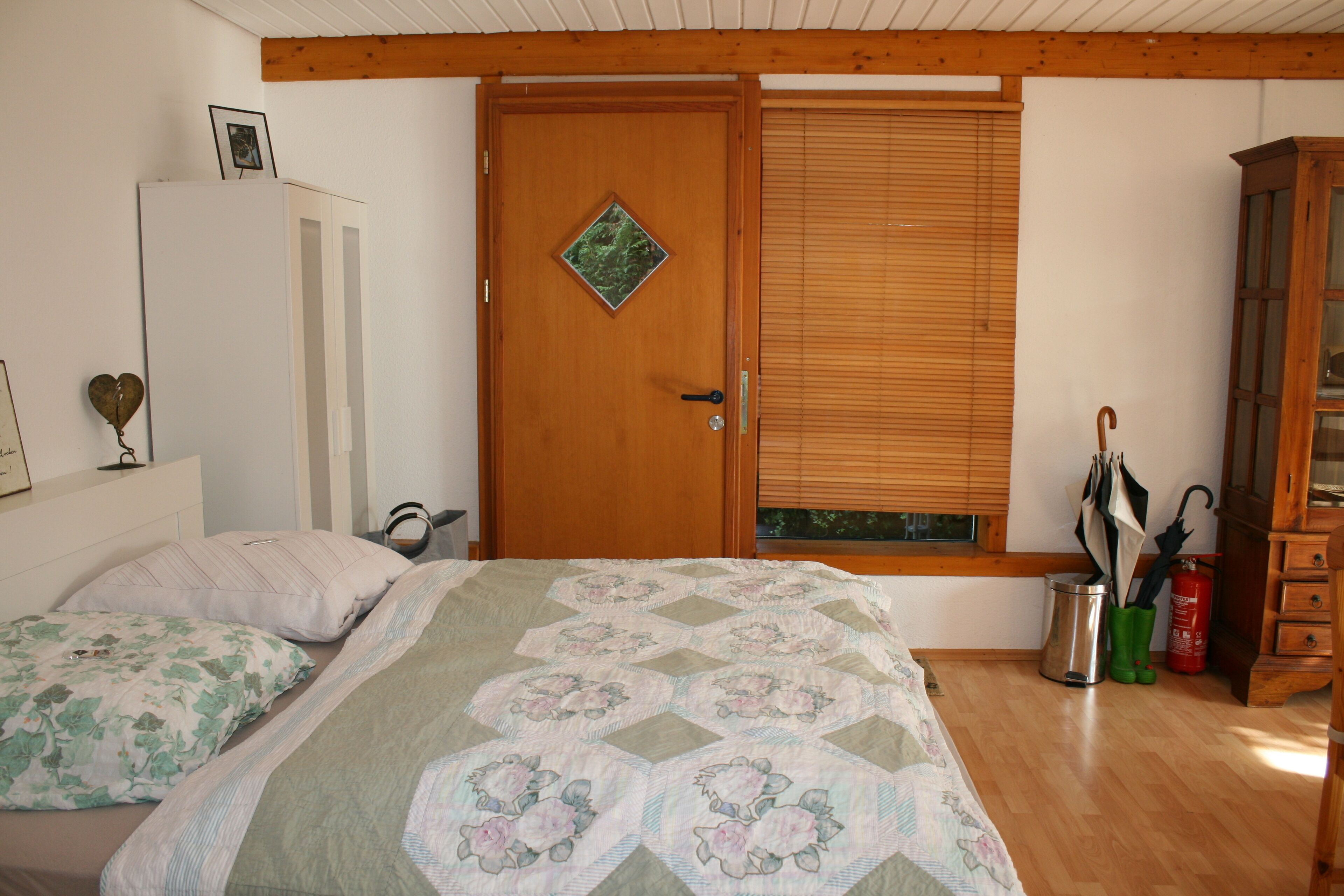 A bedroom with a wooden door, a bed with floral bedding, a bedside table, and a window with blinds.