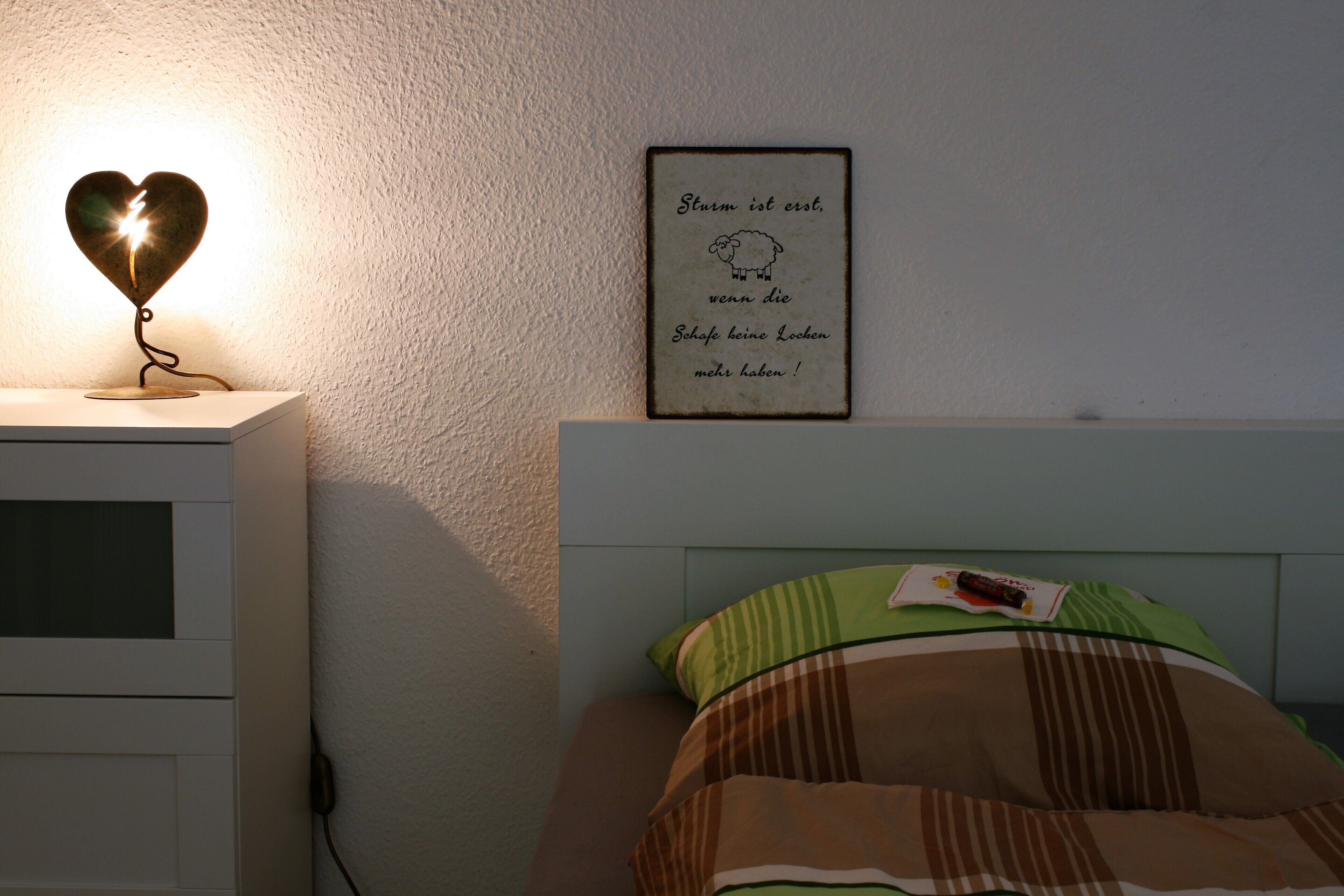 Een slaapkamer met een bedlampje, een lijst met een citaat aan de muur en een bed met een gestreept kussen.