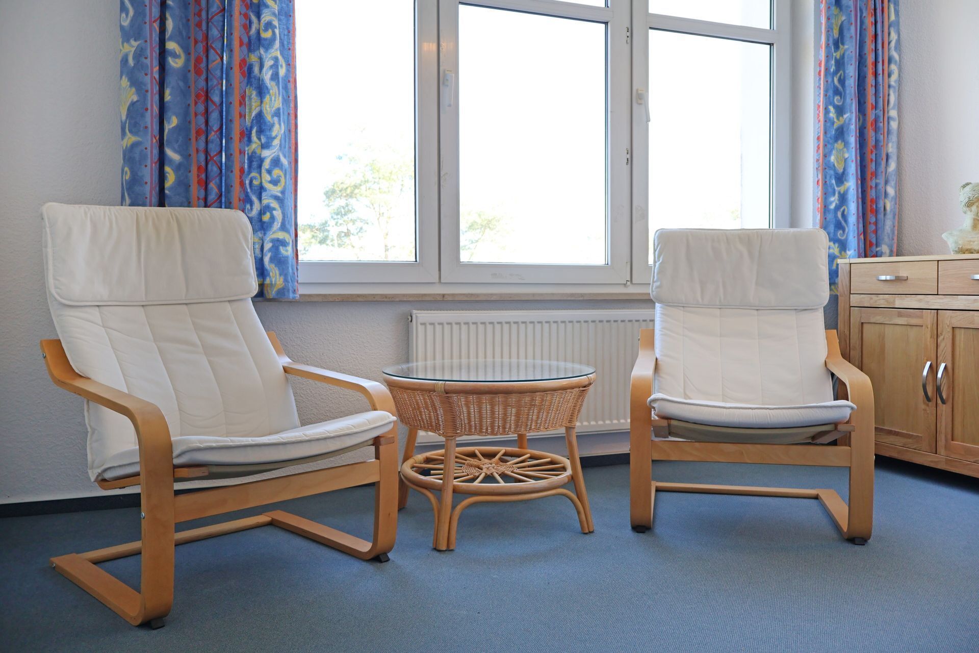 Twee witte fauteuils met houten frames, een rieten tafeltje en een uitzicht op een raam met gordijnen.