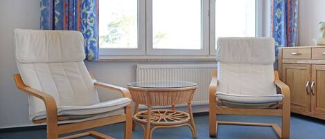Twee witte fauteuils met houten frames, een rieten tafeltje en een uitzicht op een raam met gordijnen.