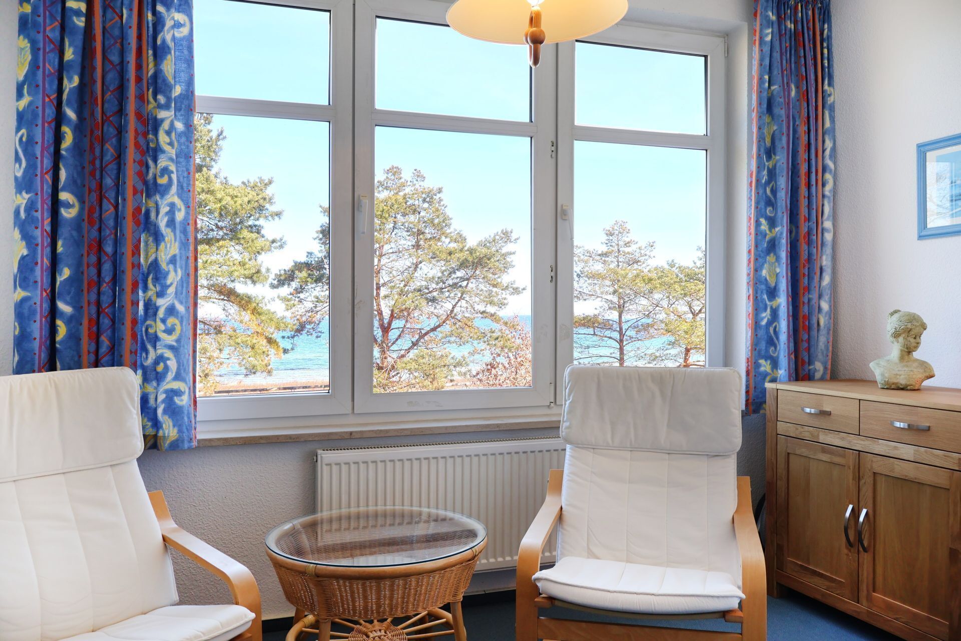 A room with two white cushioned chairs, a wicker table, a wooden cabinet, and a view of trees and water through large windows.