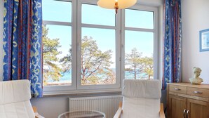 Ett rum med två vita fåtöljer, ett bord av rotting, ett träskåp och en utsikt över träd och vatten genom stora fönster.