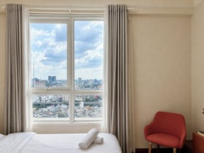 Habitación de hotel con un ventanal grande, una cama con ropa blanca, una silla roja y vistas a un paisaje urbano.