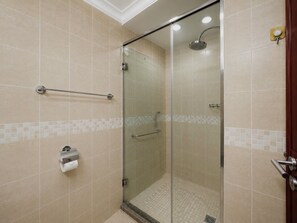 Cabina de ducha con paredes de vidrio, un cabezal de ducha manual, un perchero y un dispensador de papel higiénico.