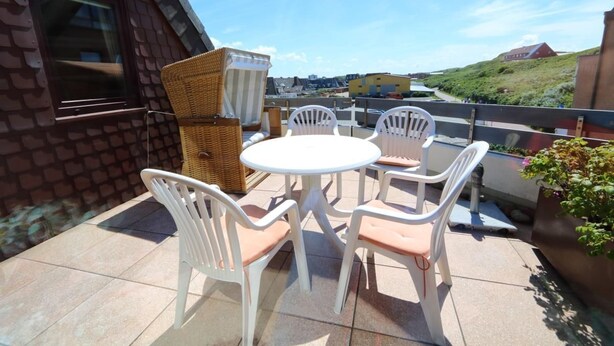 A patio with a round table and white chairs, a wicker chair, and a view of a road and buildings in the background.