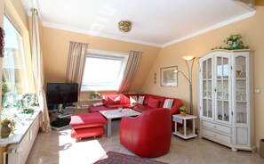 A living room with a red sofa, a television, a cabinet with glass doors, and a window with curtains. - House Alice - Block 32/13 (Sylt)