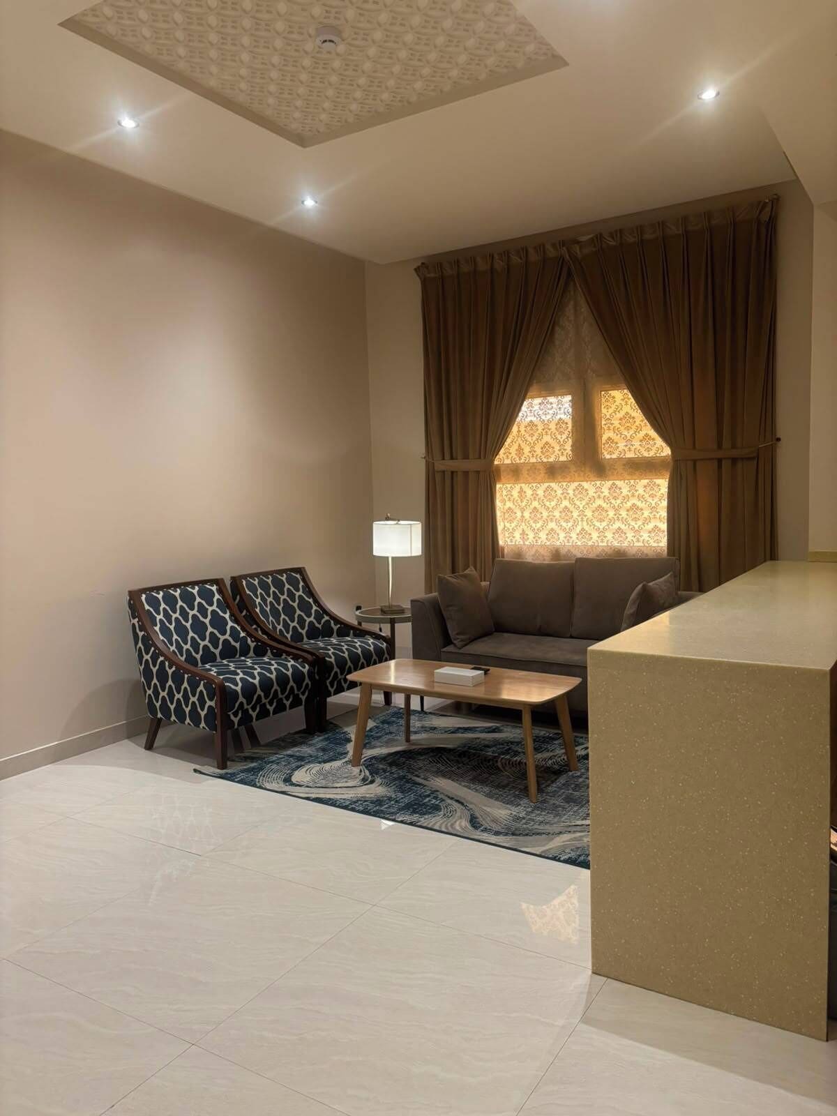 A modern living room with a sofa, a patterned armchair, a coffee table, and a large window with curtains.